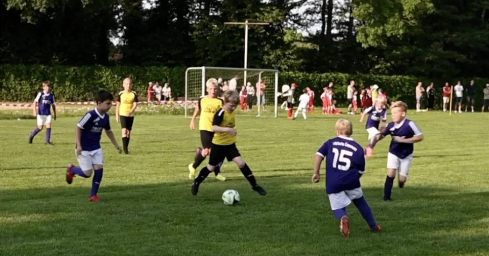 Kinder spielen Fußball auf einem grünen Rasen; gelbe und blaue Trikots, Zuschauer im Hintergrund.