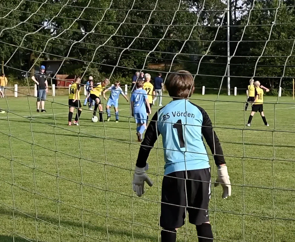 Jugendfußballspiel: Torwart in blauem Trikot konzentriert, Spieler in gelb und blau auf grünem Rasen.