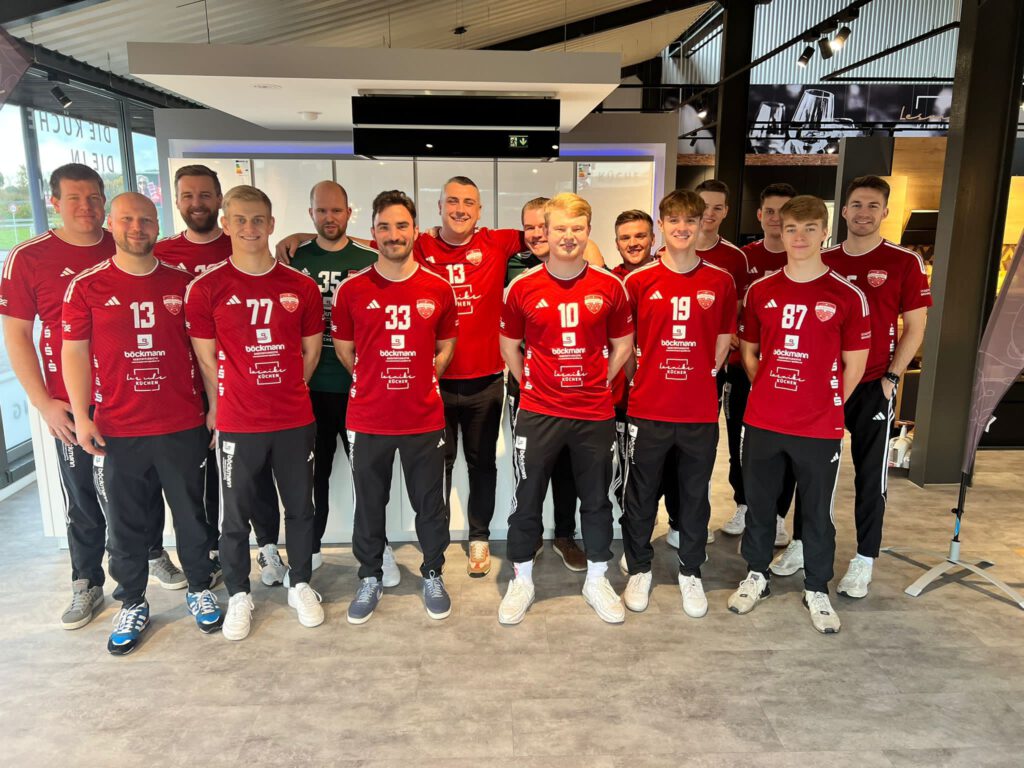 Eine Gruppe von Personen in roten Sporttrikots posiert lächelnd für ein Teamfoto in einem modernen Raum.