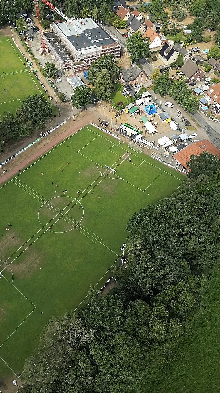 Luftaufnahme eines Fußballspiels neben einer Baustelle, umgeben von Wohnhäusern und parkenden Autos.
