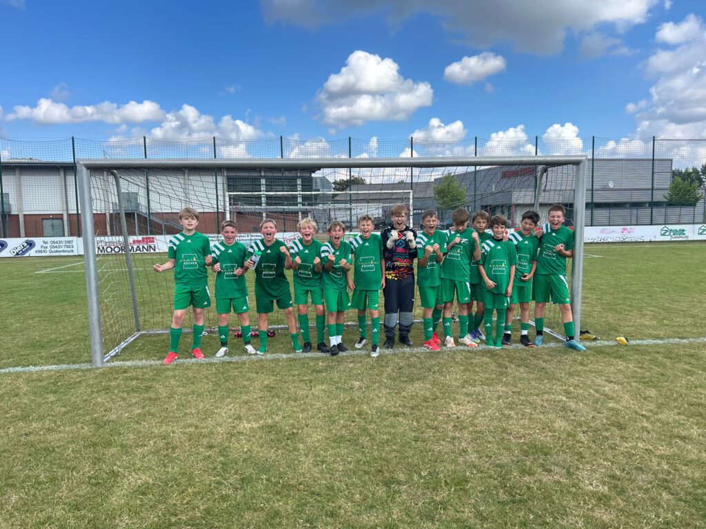 Kinder in grünen Fußballtrikots jubeln vor einem Tor auf einem sonnigen Sportplatz.