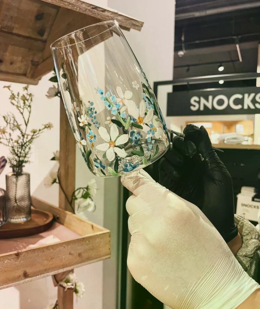 Ein kunstvoll bemaltes Weinglas wird von behandschuhten Händen in einem kreativen Atelier gehalten.