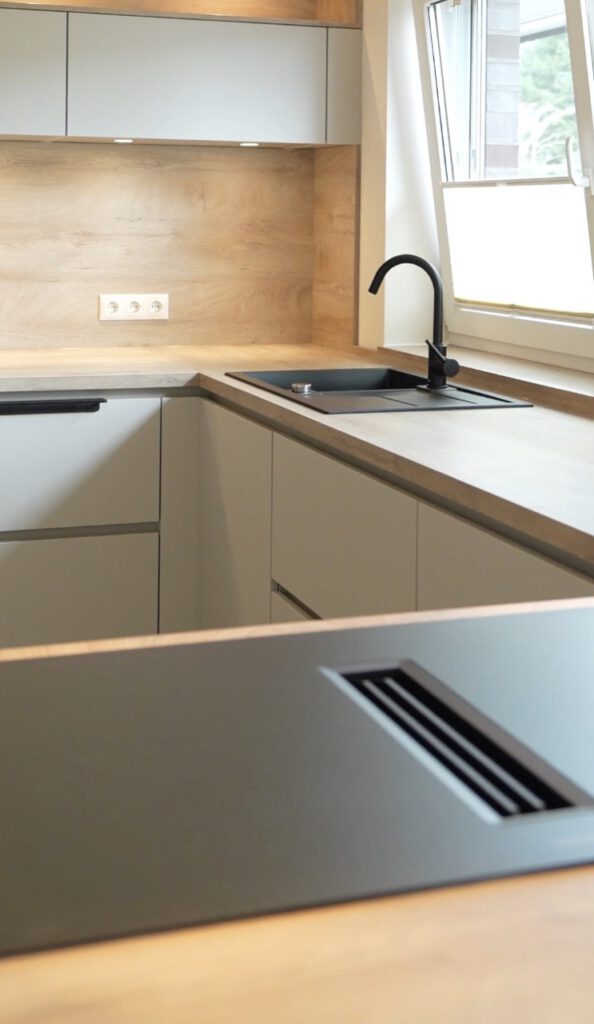 Moderne Küche mit hellgrauen Schränken, Holzoptik-Arbeitsplatte, schwarzem Spülbecken und Tageslicht.