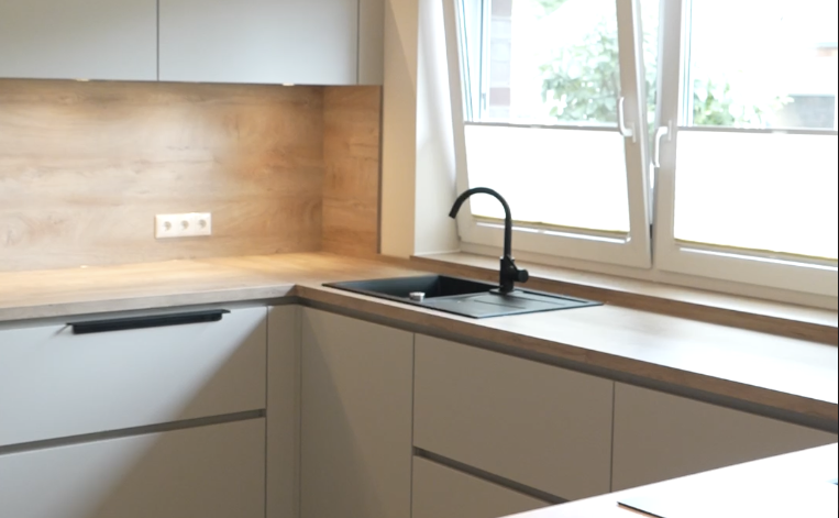 Moderne, helle Küche mit grauen Schränken, Holzarbeitsplatte und natürlichem Licht durch ein Fenster.
