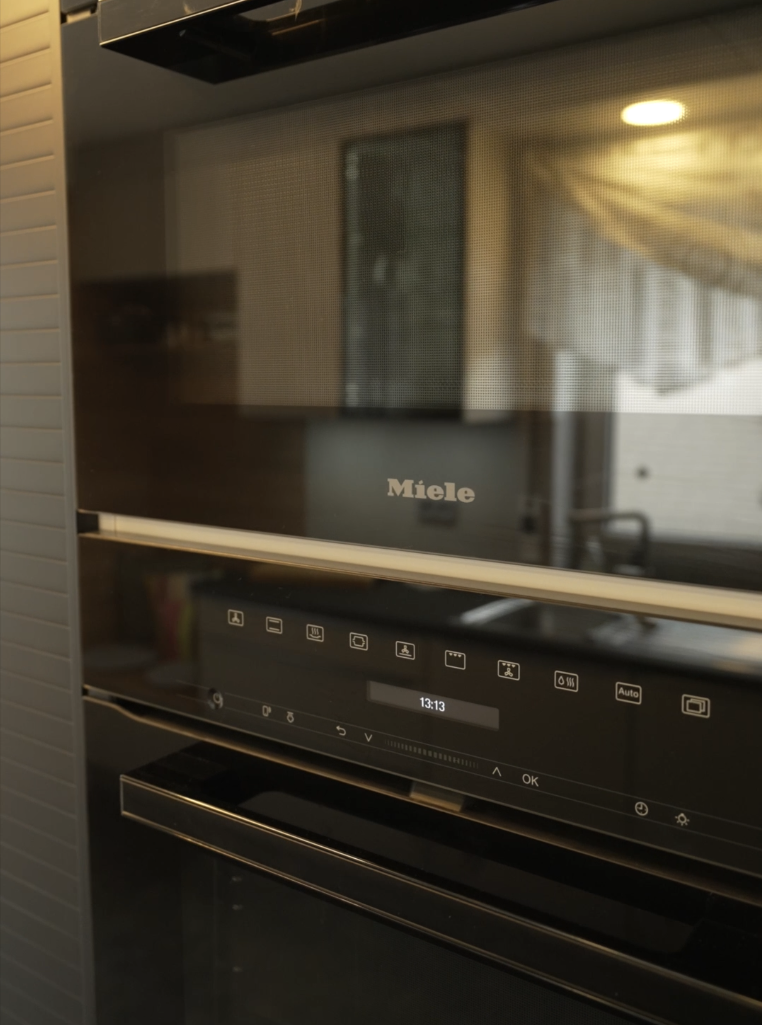 Moderner Miele-Backofen mit schwarzer Glasfront, zeigt Uhrzeit 13:13, spiegelt warmen Raum wider.