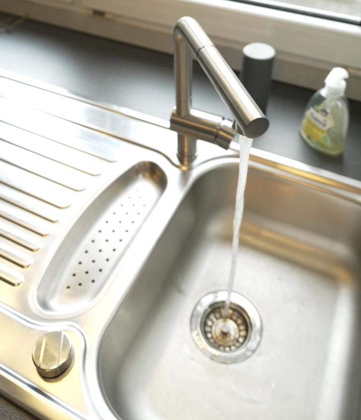 Moderne Edelstahlspüle mit gebogenem Wasserhahn, aus dem Wasser fließt; grüner Seifenspender im Hintergrund.