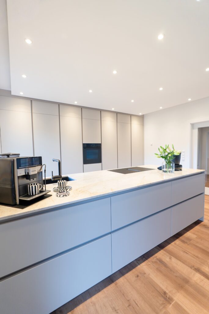 Moderne Küche mit Marmorinsel, Kaffeevollautomat, Pflanzenvase und glatten Schränken in neutralen Tönen.
