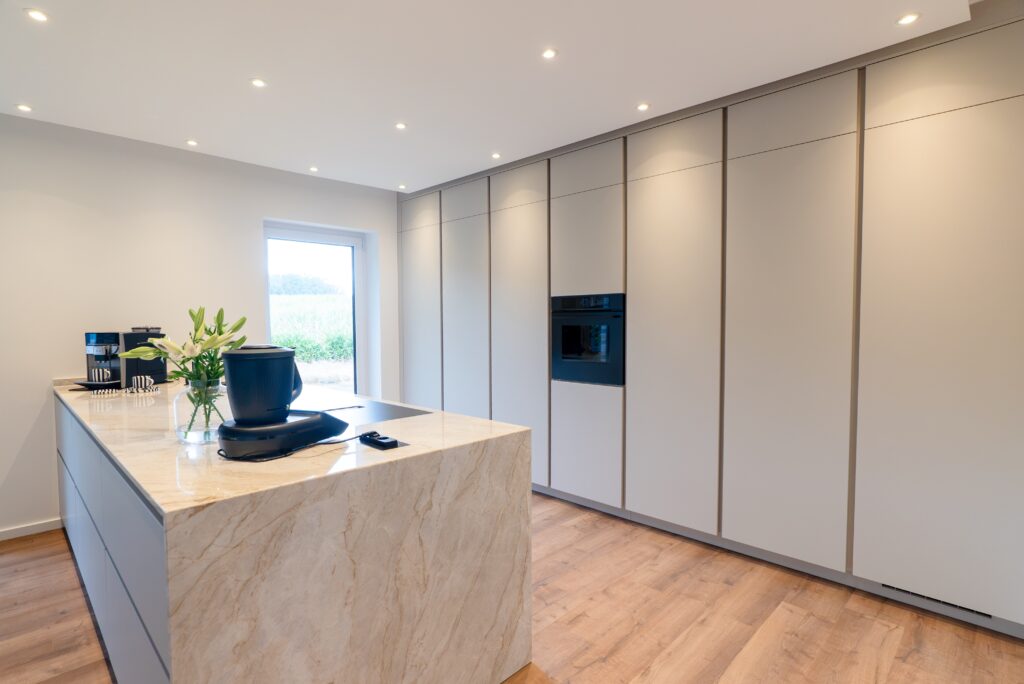 Moderne, minimalistische Küche mit heller Insel, Blumenstrauß und eingebautem Ofen, umgeben von Grün.