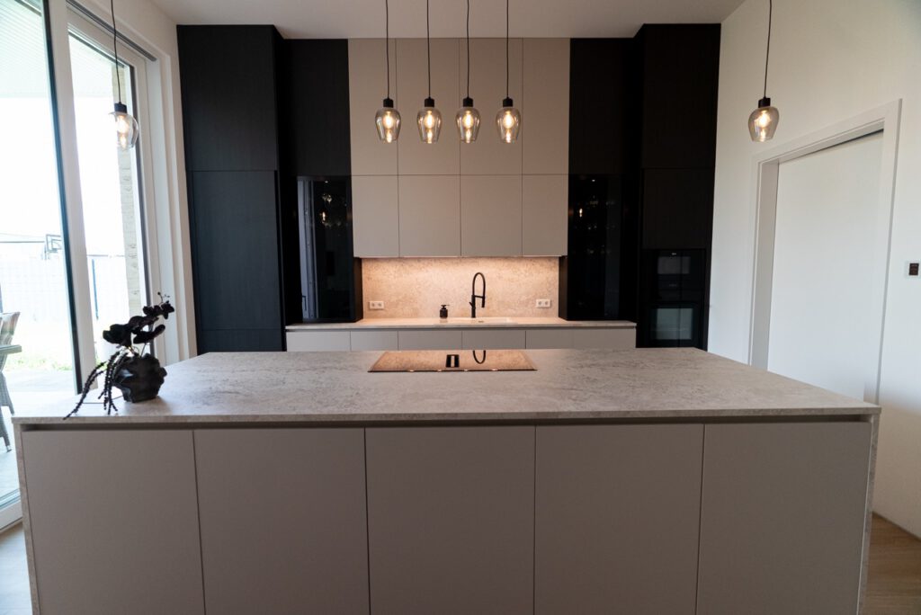 Moderne Küche mit heller Insel, Kochfeld, Spüle, hängenden Lampen und dekorativer Pflanze.