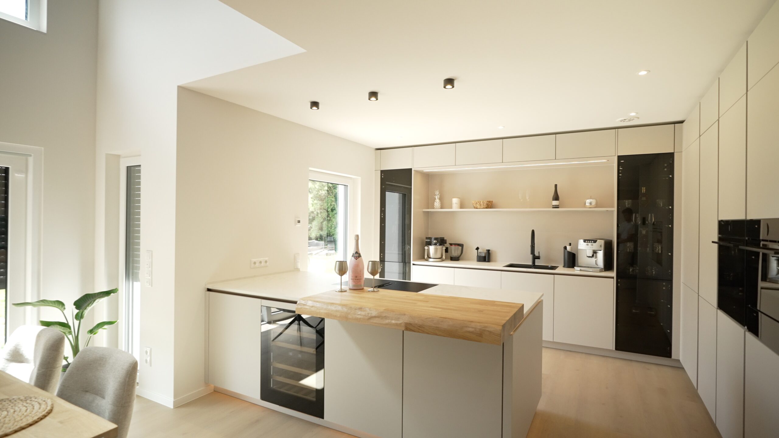 Moderne, minimalistische Küche mit heller Holzinsel, Weingläsern, Einbaugeräten und eleganten Details.