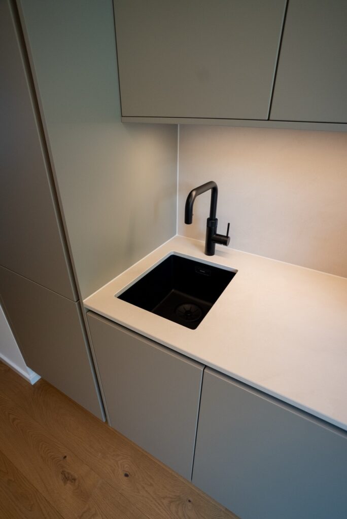 Moderne Küchenecke mit hellen Schränken, schwarzem Wasserhahn und Holzfußboden in minimalistischem Stil.