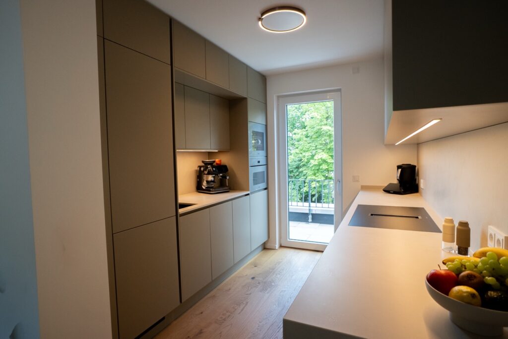 Moderne, minimalistische Küche mit beigen Schränken, Obstschale und Blick auf grüne Bäume durch Glastür.