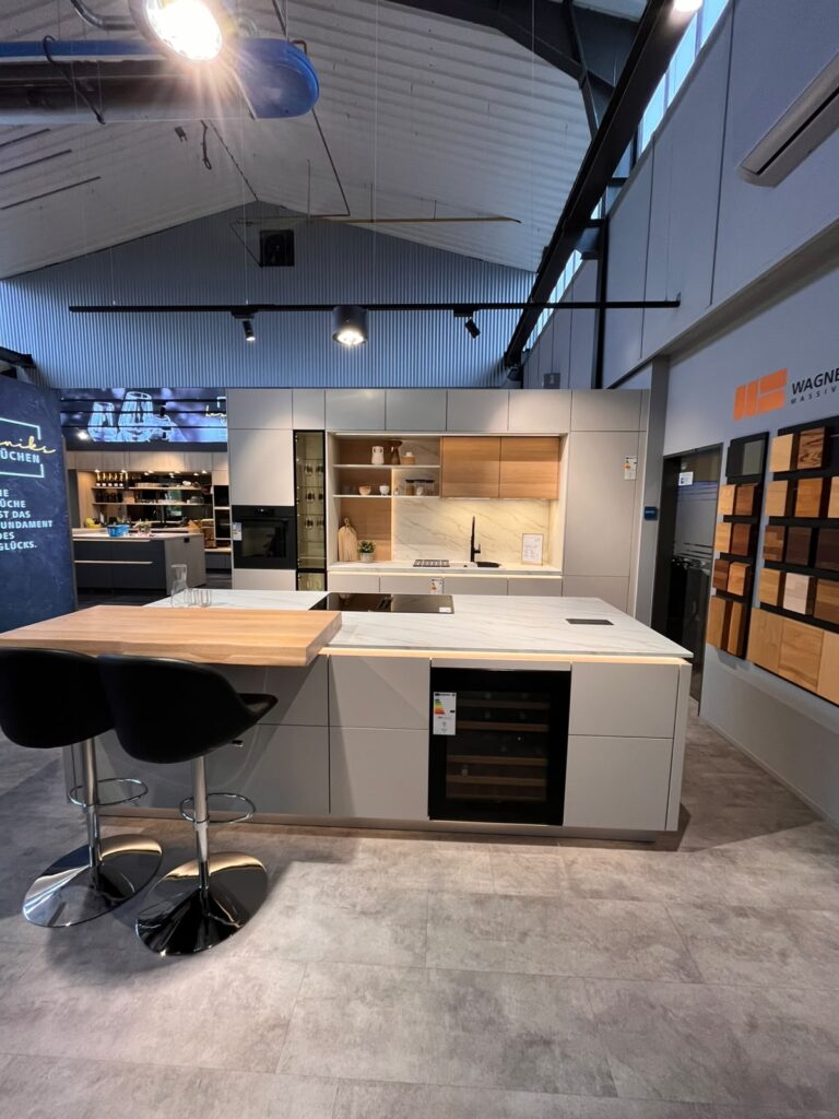 Moderne Küchenausstellung mit heller Insel, schwarzen Barhockern und integriertem Weinkühlschrank.