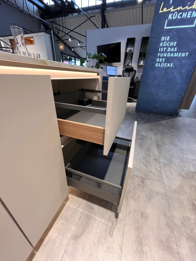 Moderne Küchenausstellung mit offenen Schubladen, neutralen Farben und einem Werbebildschirm.