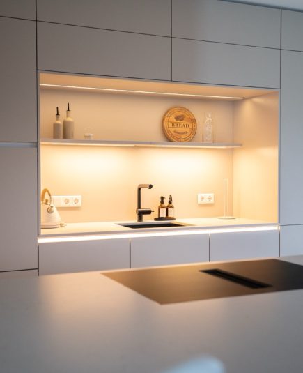 Moderne, minimalistische Küche mit hellen Fronten, schwarzer Armatur und dekorativ beleuchtetem Regal.