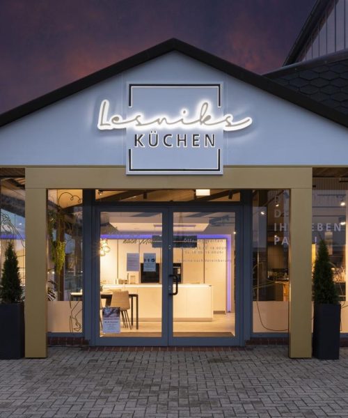 Moderne Küchenstudio-Fassade mit Glasfront, leuchtendem „Lesniks KÜCHEN“-Schild und Pflanzen.