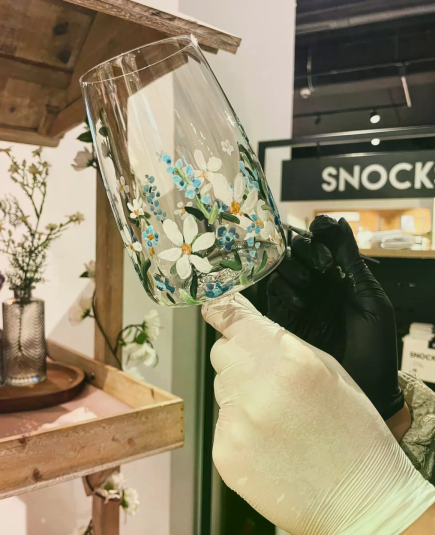 Ein kunstvoll bemaltes Weinglas wird von behandschuhten Händen in einem kreativen Atelier gehalten.